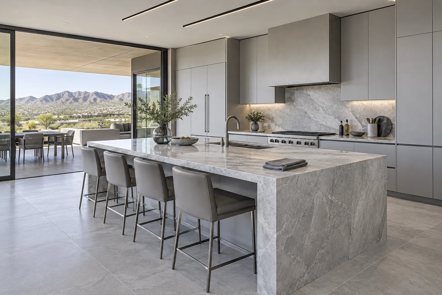 Pale grey kitchen with a long marble island and stools, sliding glass walls open to a patio and the desert mountains